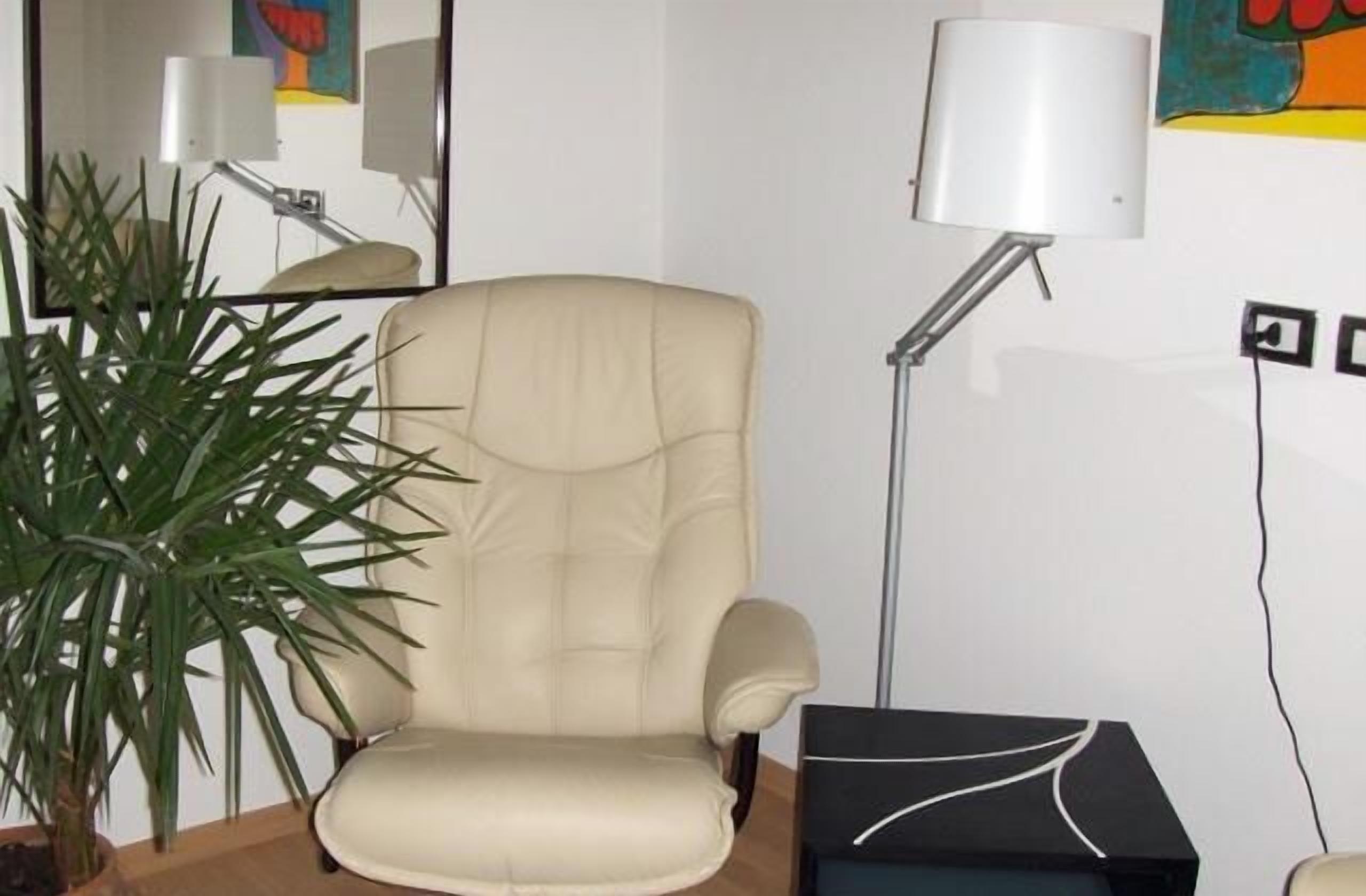 A beige leather chair, a floor lamp, a black box, and a potted plant in a room with a white wall and a colorful painting.