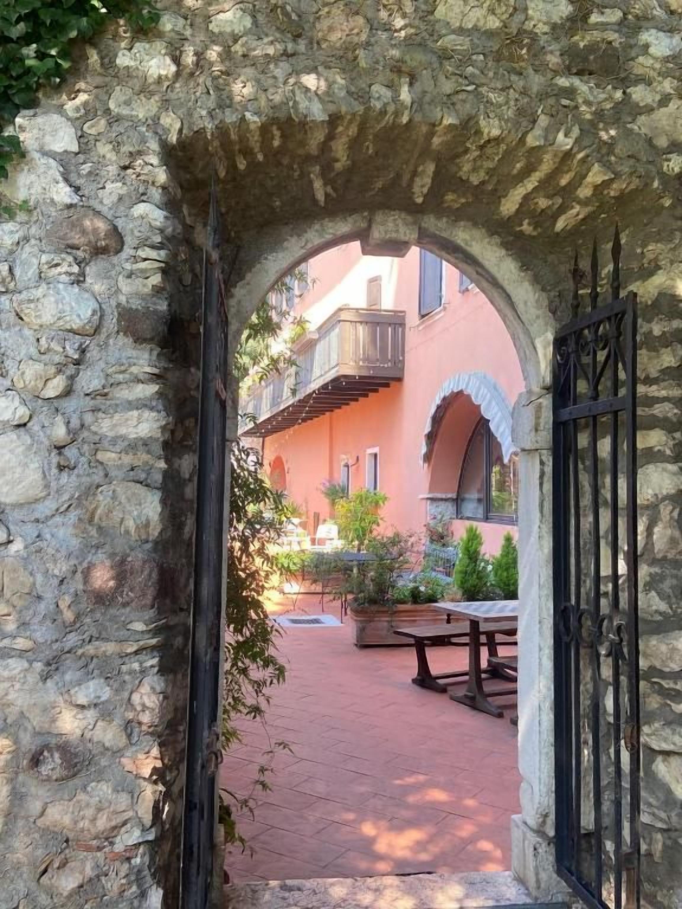An arched stone doorway leading to a courtyard with a table and chairs, a building with balconies, and a gate.