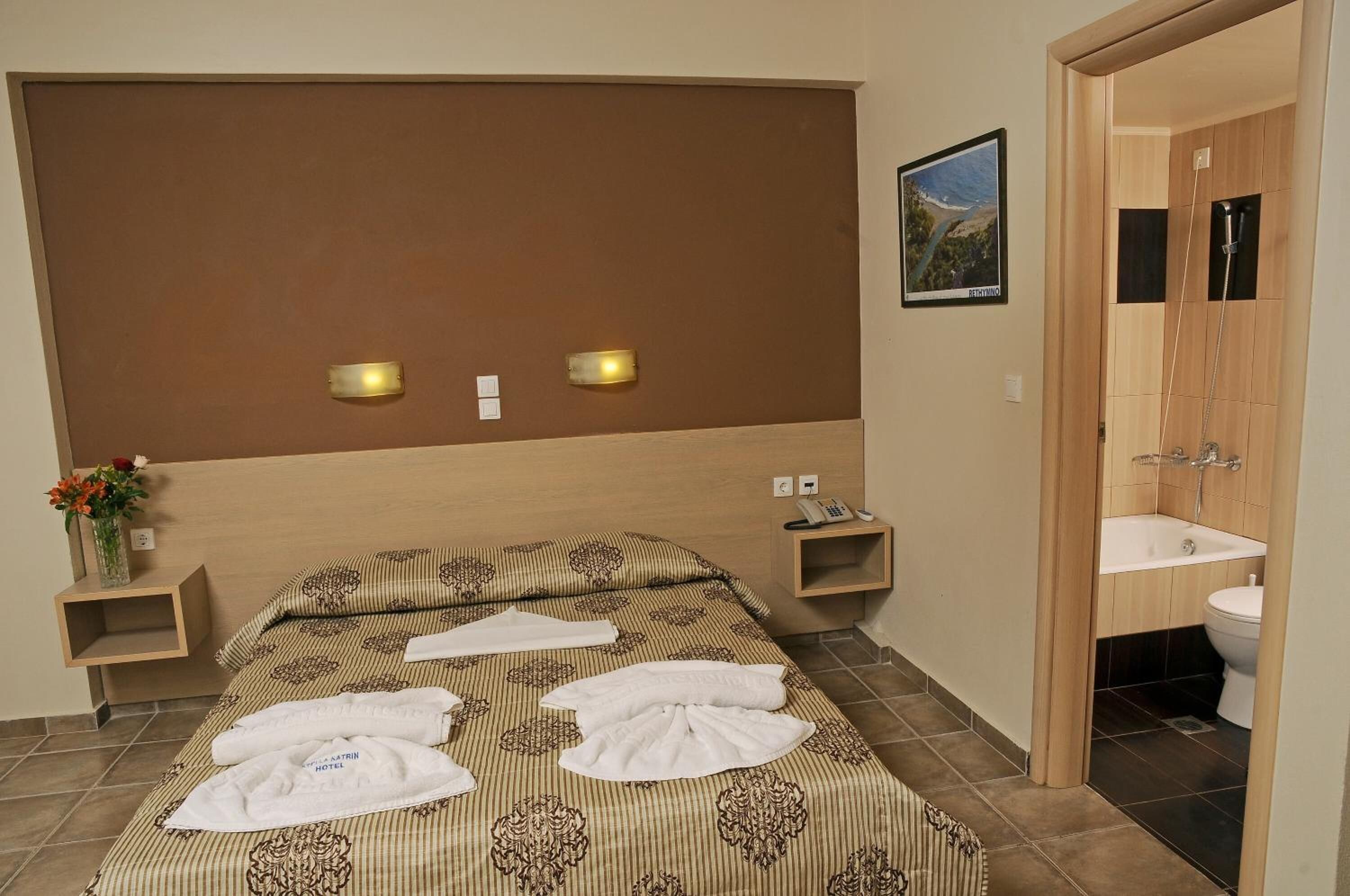 A hotel room with a bed, bedside tables, a nightstand, a bathroom with a sink and toilet, and a framed picture on the wall.