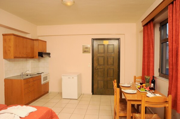 A compact kitchen with wooden cabinets, a microwave, and a small refrigerator. There is a dining area with a table set for two, a bed with a red cover, and a door with a sign.