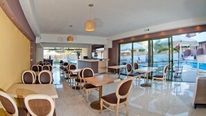A hotel dining area with tables and chairs, a reception desk, and a pool area visible through large windows. - Hotel Stella Katrin (Rethymno)