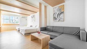 Quarto de hotel moderno com sofá cinza, mesa de centro de madeira, cama com roupa de cama branca e uma grande pintura abstrata na parede.