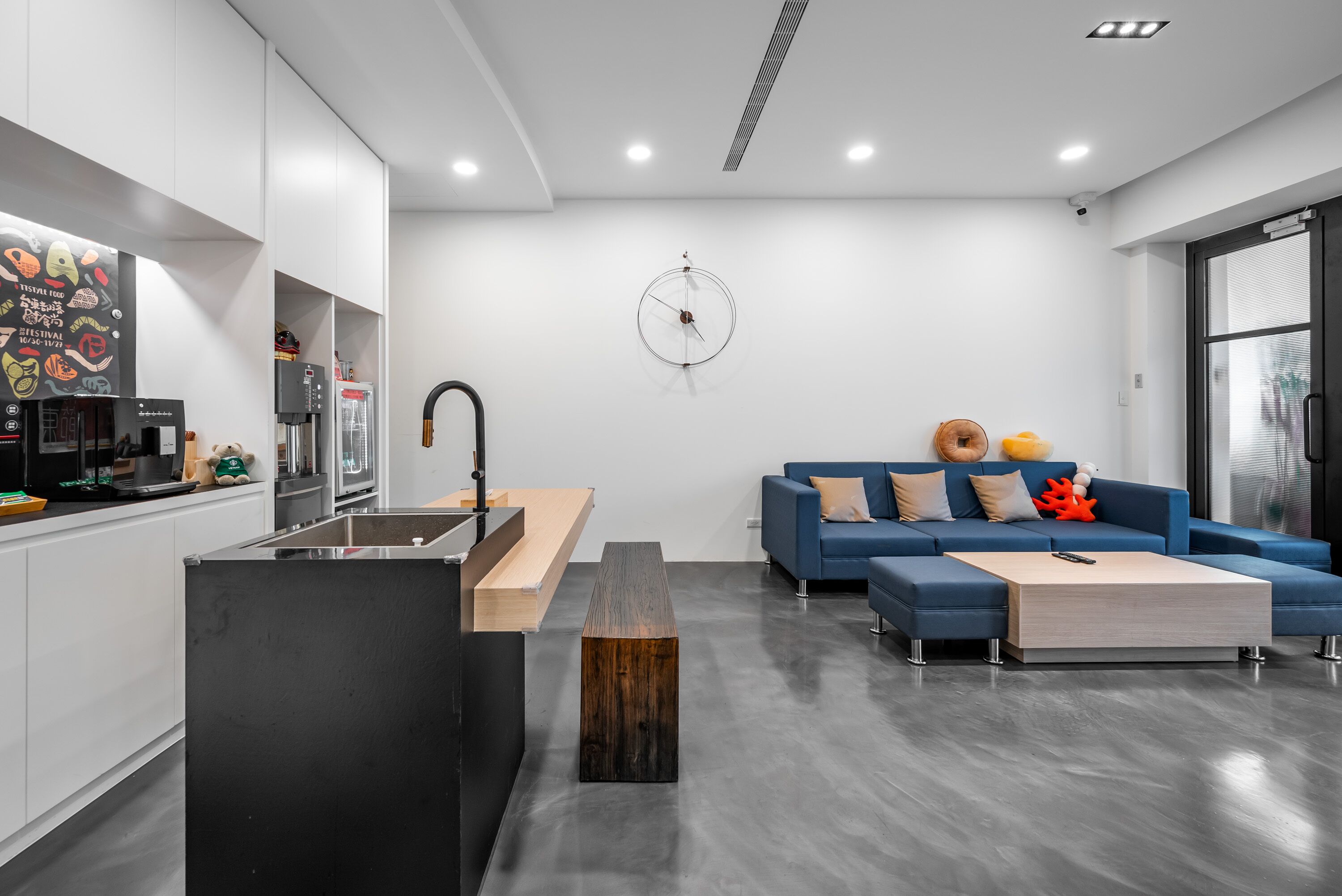 A modern kitchen with a built-in coffee machine, a sink, and a wooden island. There is a blue sofa with decorative pillows and a coffee table in the living area.