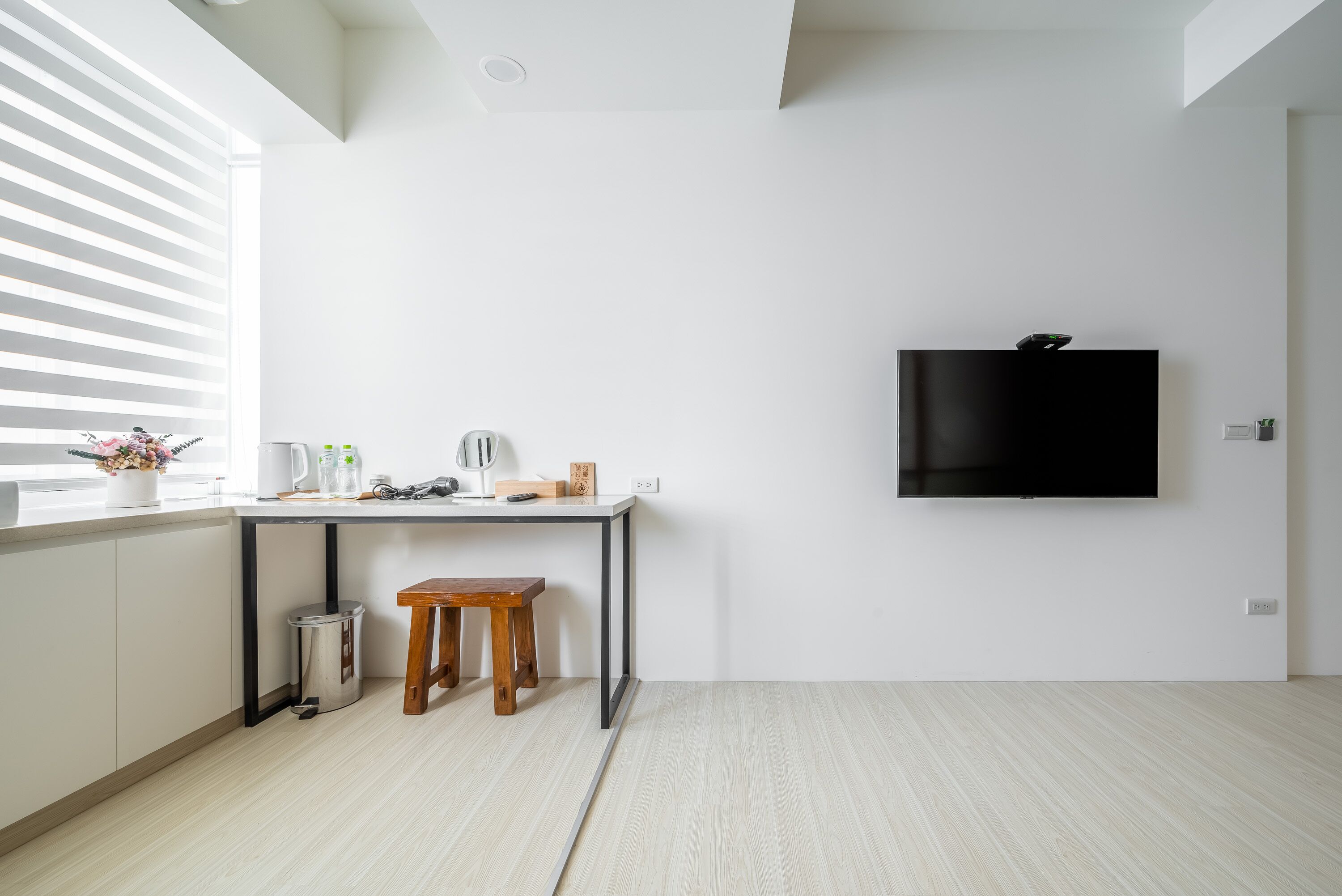 A modern kitchen with a white countertop, a wooden stool, a trash bin, and a wall-mounted TV.