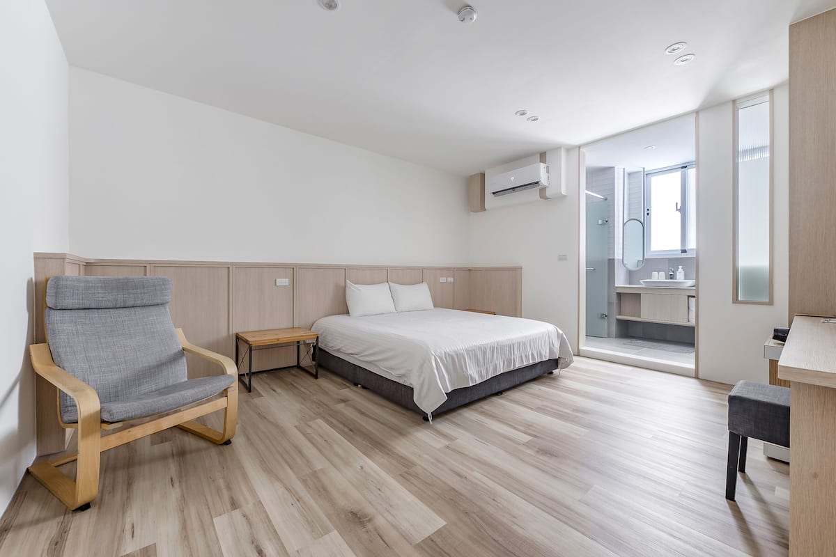 Ein modernes Schlafzimmer mit einem Bett, einem Sessel, einem Schreibtisch und einem durch eine offene Tür einsehbarer Badezimmer.