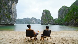 Two people seated on beach chairs facing a calm sea with rocky cliffs in the background. - Heritage Line Violet cruise – Ha Long Bay & Lan Ha Bay  (Tuan Chau)