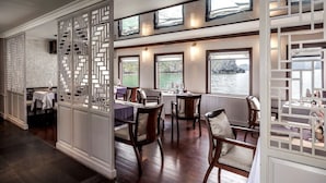 A dining area with wooden tables and chairs, white decorative screens, and large windows overlooking a body of water. - Heritage Line Violet cruise – Ha Long Bay & Lan Ha Bay  (Tuan Chau)