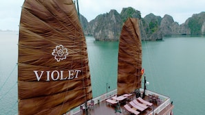A ship with large brown sails and the word "VIOLET" on one of them. The ship has a deck with lounge chairs and a railing. - Heritage Line Violet cruise – Ha Long Bay & Lan Ha Bay  (Tuan Chau)