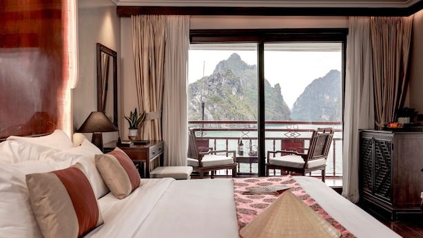 A hotel room with a large bed, a balcony with a view of mountains, a desk with a lamp, and a chair. - Heritage Line Violet cruise – Ha Long Bay & Lan Ha Bay  (Tuan Chau)