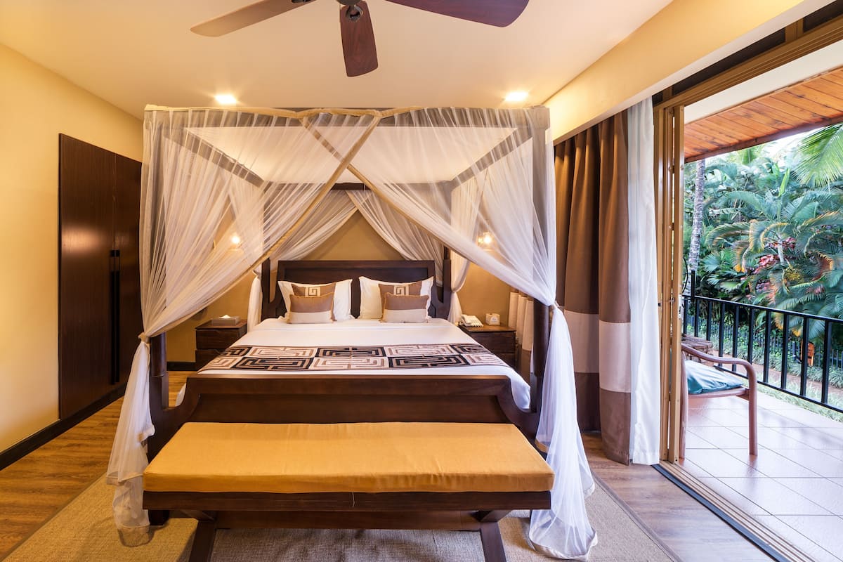 A bedroom with a four-poster bed, a bench, a nightstand, and a view of the outdoors.