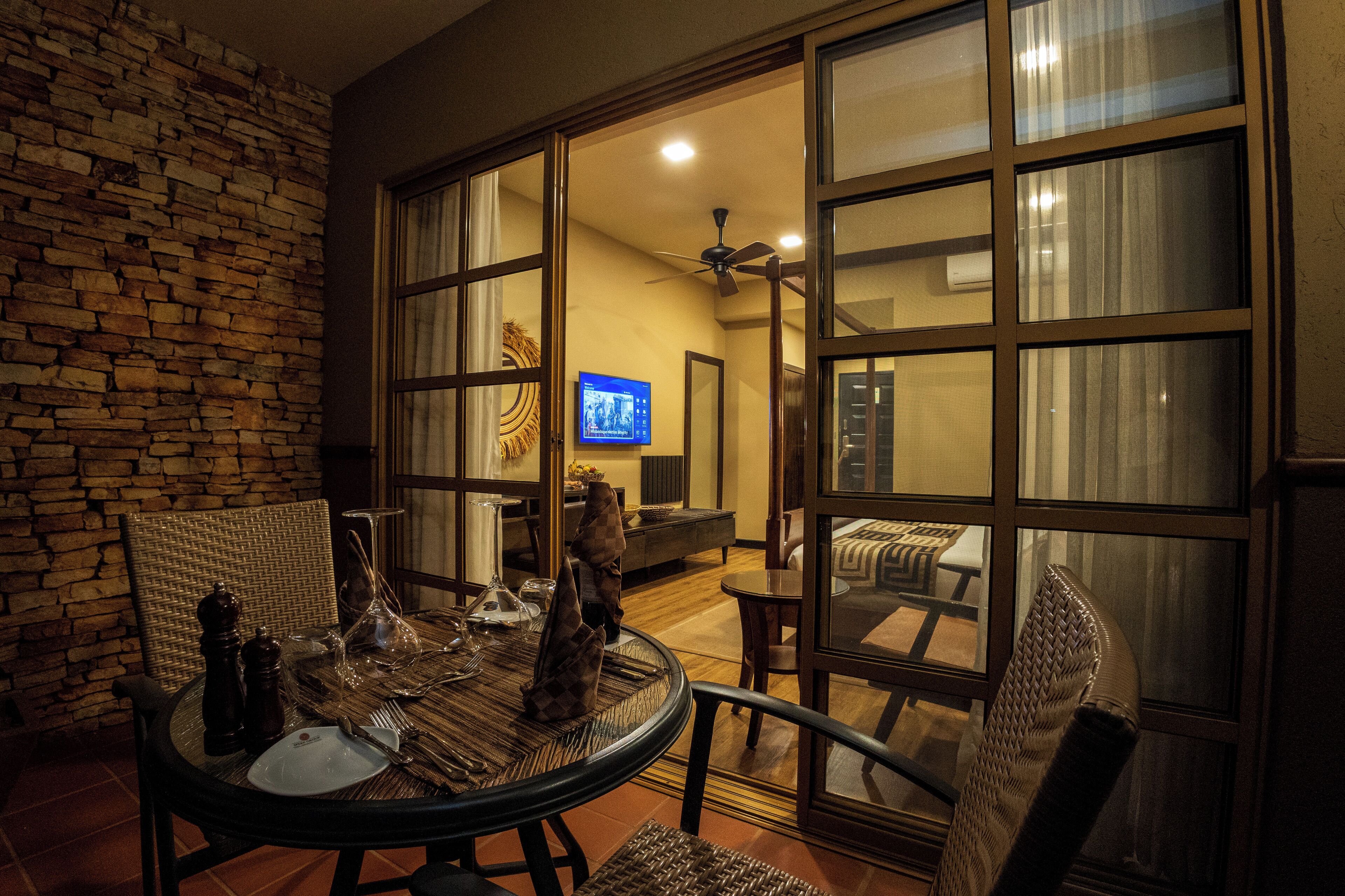 A dining area with a table set for two, a wicker chair, and a view through a glass door into a living room with a TV and a ceiling fan.