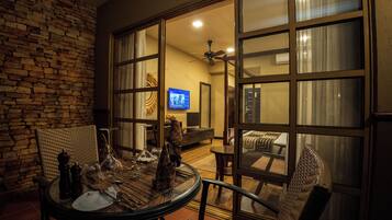 A dining area with a table set for two, a wicker chair, and a view through a glass door into a living room with a TV and a ceiling fan.