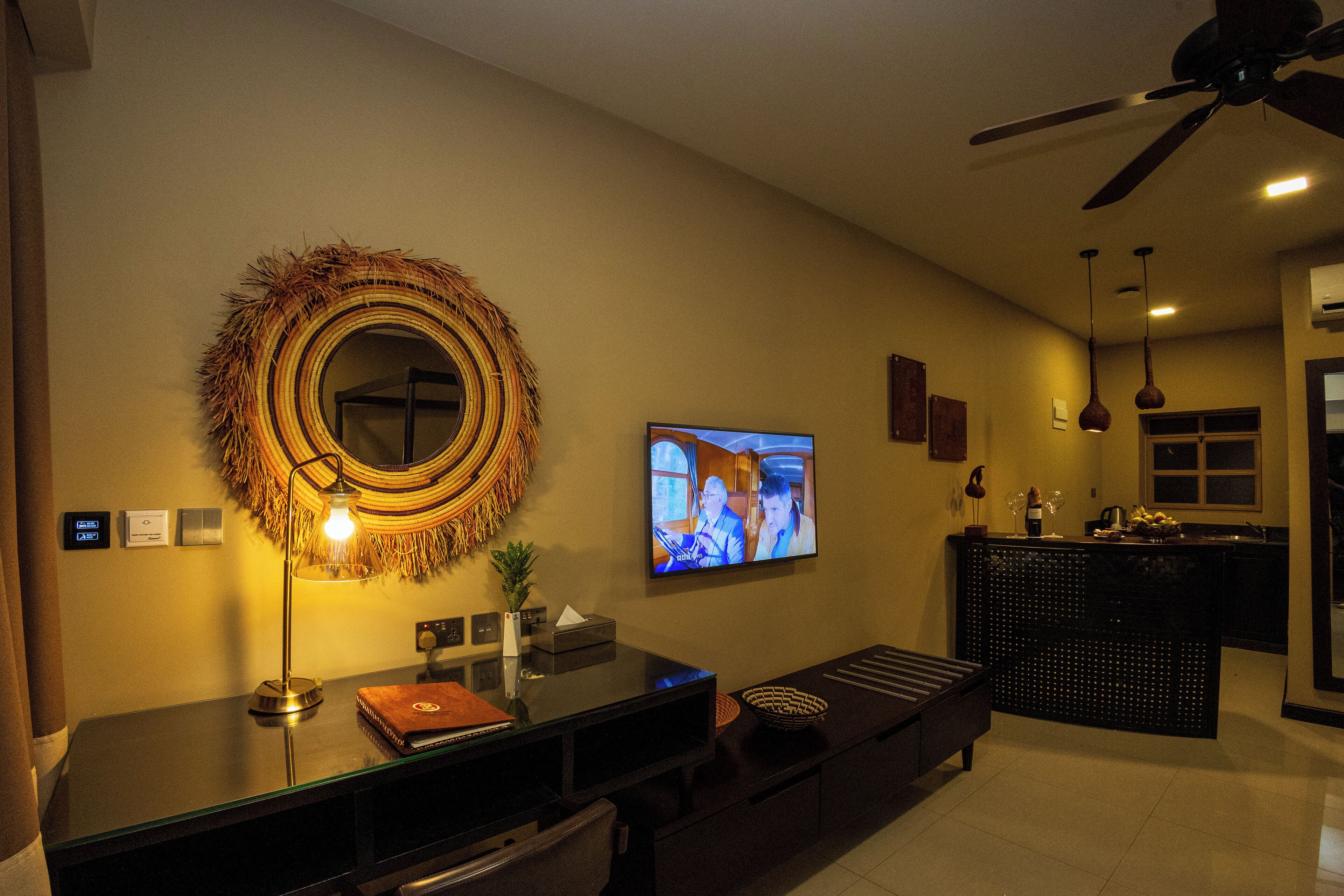 A modern living room with a television, a decorative wall hanging, a desk with a lamp, and a ceiling fan.