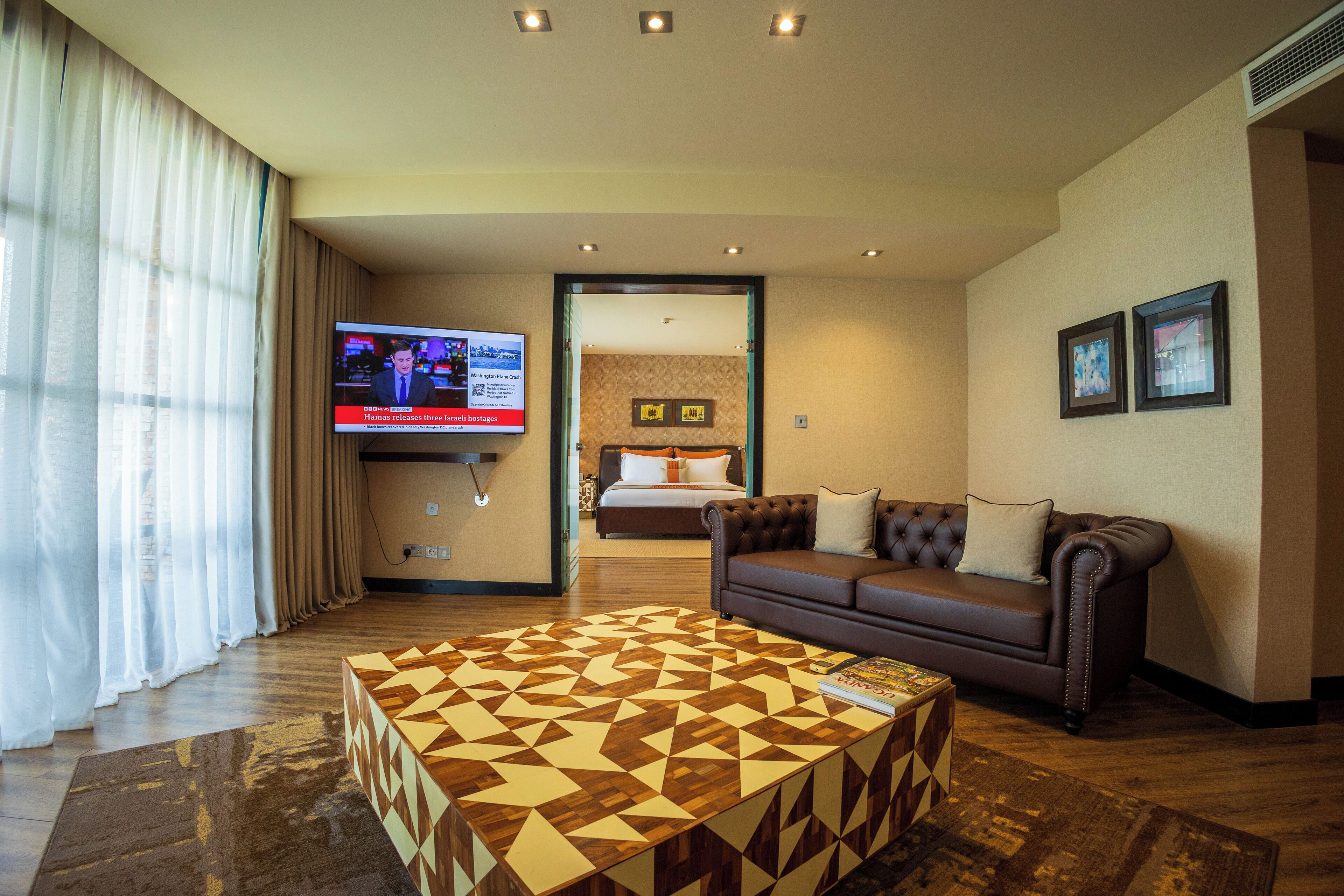 A hotel room with a large TV, a brown leather sofa, a patterned coffee table, and a bed with white and orange bedding.