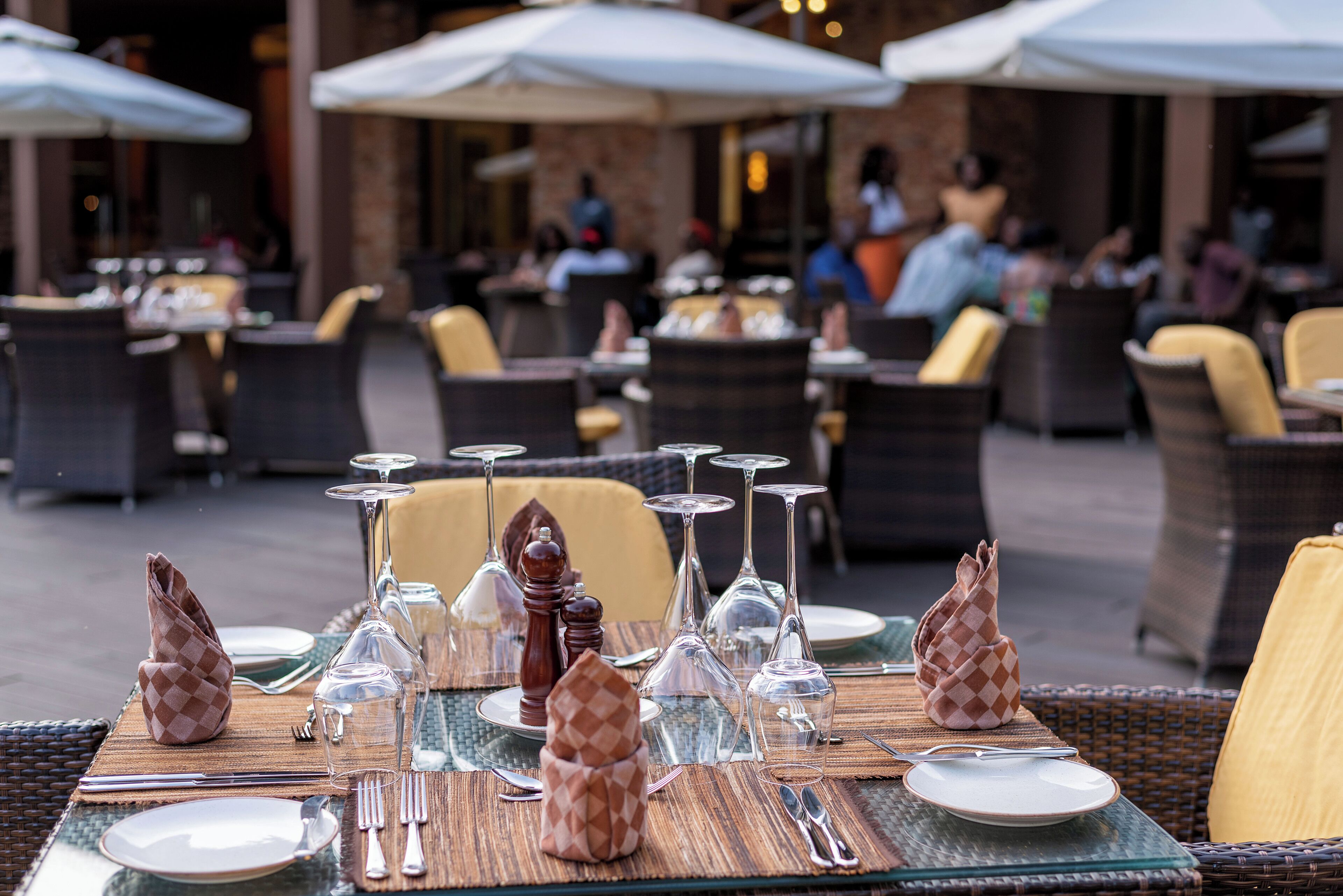 An outdoor dining area with wicker chairs and tables set for a meal, featuring clear glassware and neatly arranged cutlery.