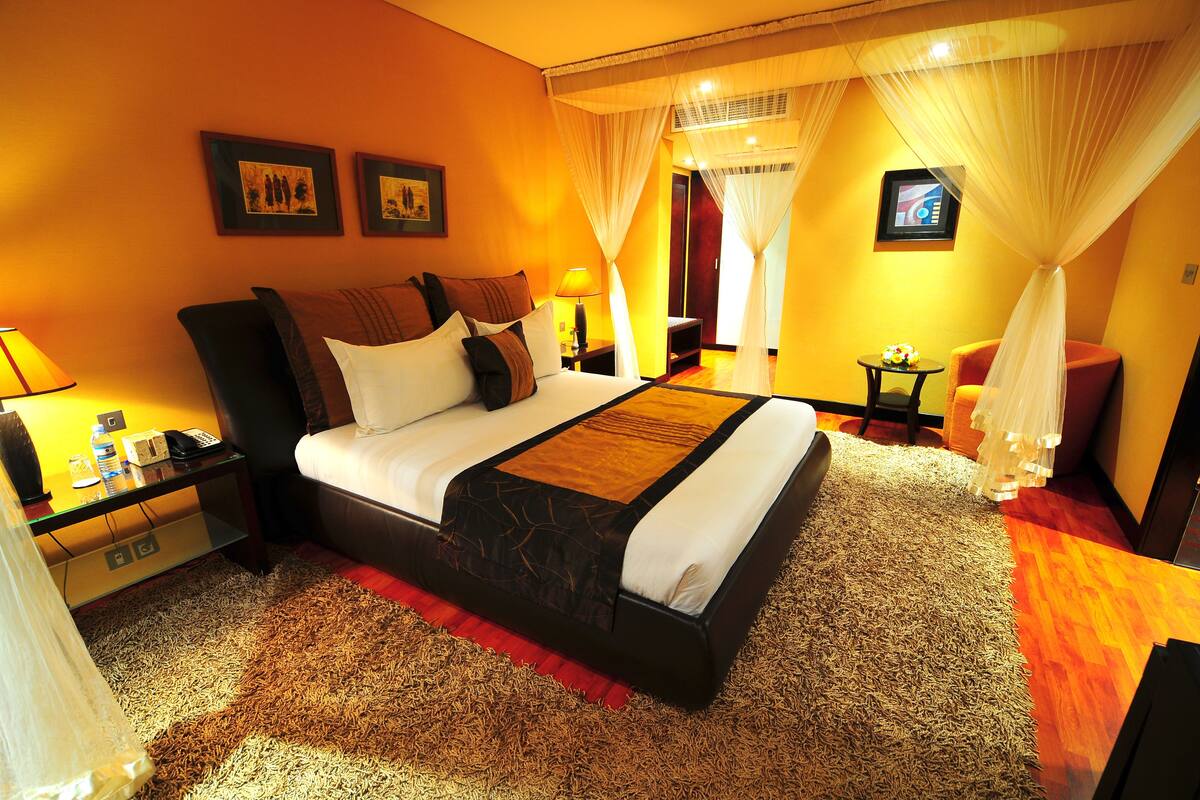 A hotel room with a large bed, a bedside table with a lamp, a small table with a vase, and a chair.