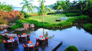 A serene outdoor dining area with tables and chairs on a wooden deck, overlooking a calm pond and lush greenery. - Speke Resort & Conference Centre (Kampala)