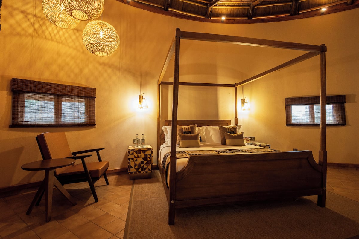 A four-poster bed with a wooden frame, a small round table, and two chairs. There are two wall-mounted lamps and a window with blinds.