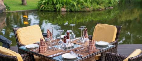 A dining table set with chairs, surrounded by a pond and greenery.