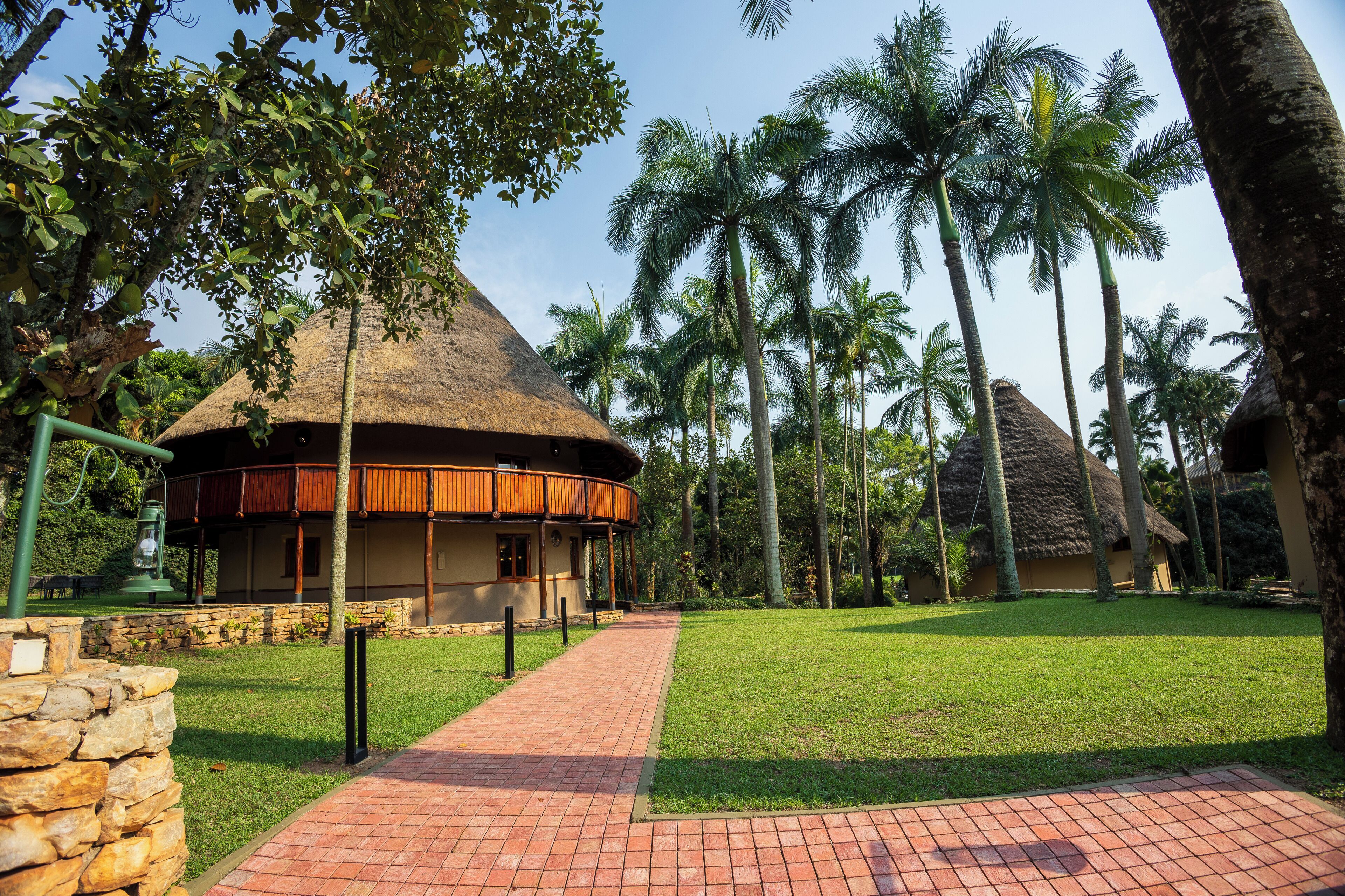 A thatched-roof building with a wooden balcony, surrounded by palm trees and a paved walkway.