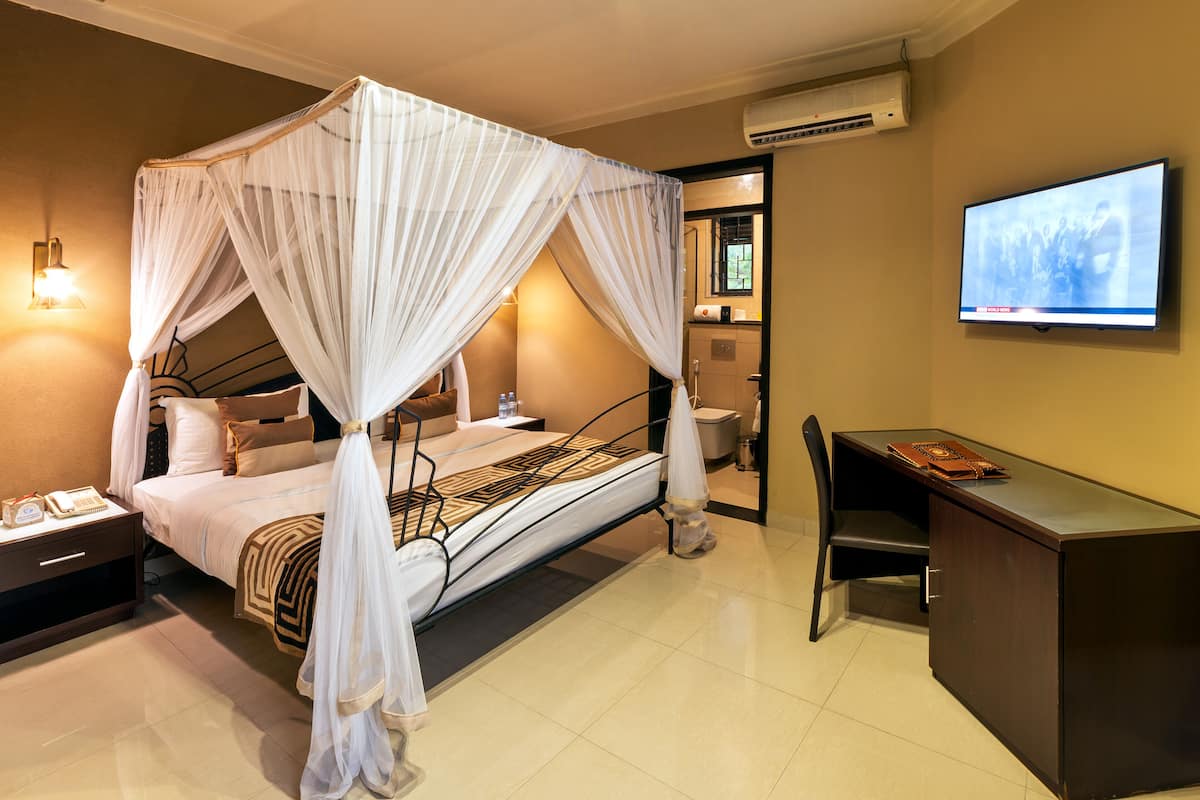 A hotel room with a four-poster bed, a desk, a chair, a television, and a bedside table with a lamp.