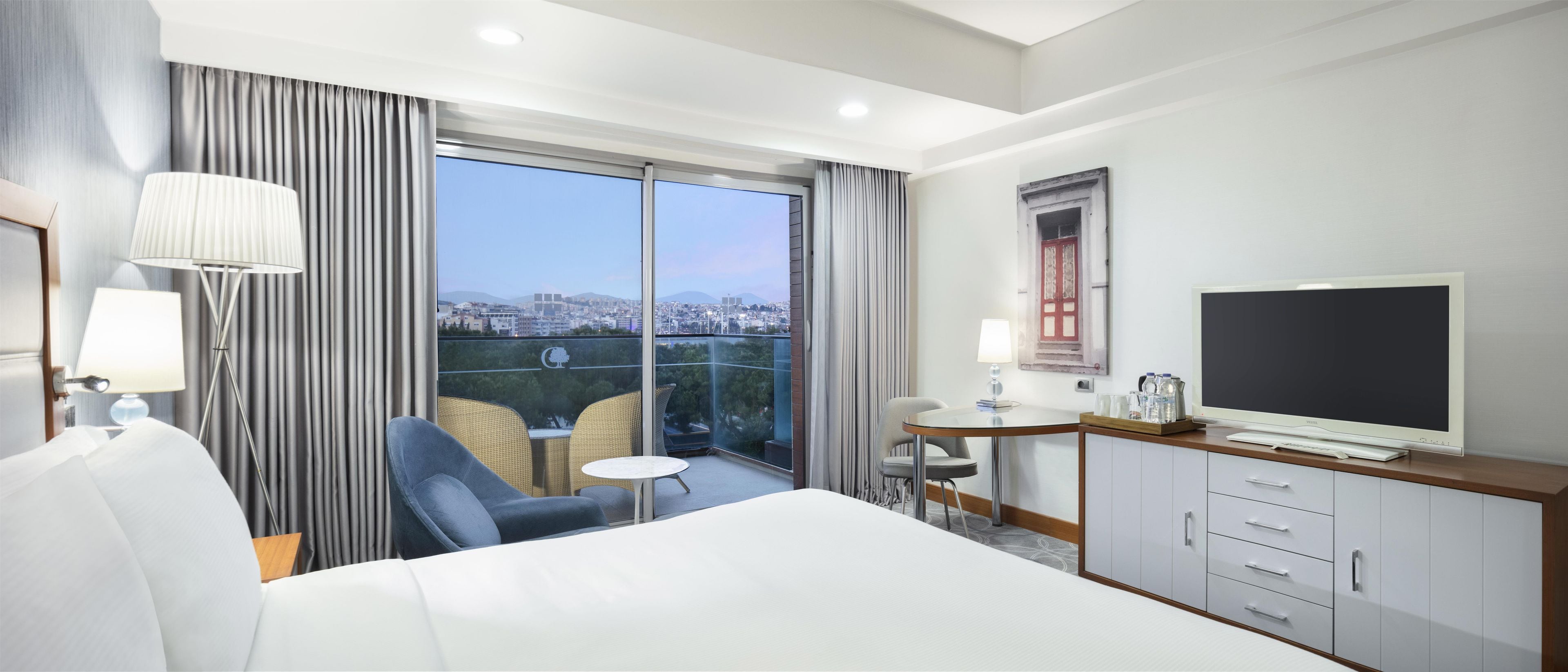 A hotel room with a bed, a TV, a desk with a chair, and a balcony with a view of the city.
