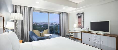 A hotel room with a bed, a TV, a desk with a chair, and a balcony with a view of the city.