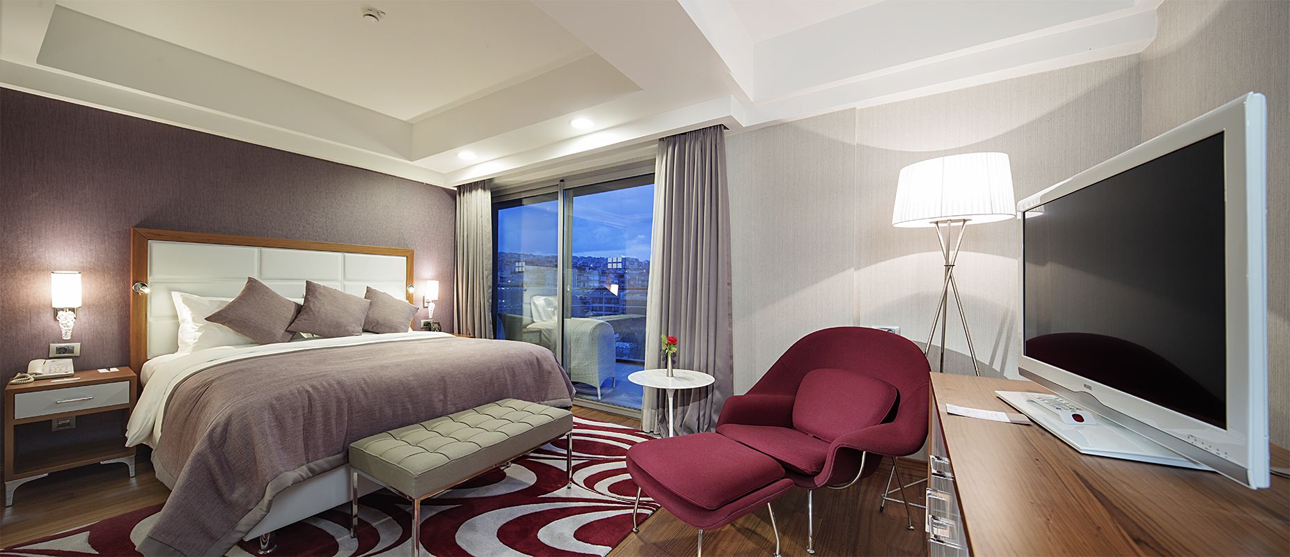 A hotel room with a bed, a desk with a computer, a chair, a bedside table, a lamp, and a view of the city.