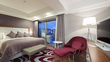 A hotel room with a bed, a desk with a computer, a chair, a bedside table, a lamp, and a view of the city.