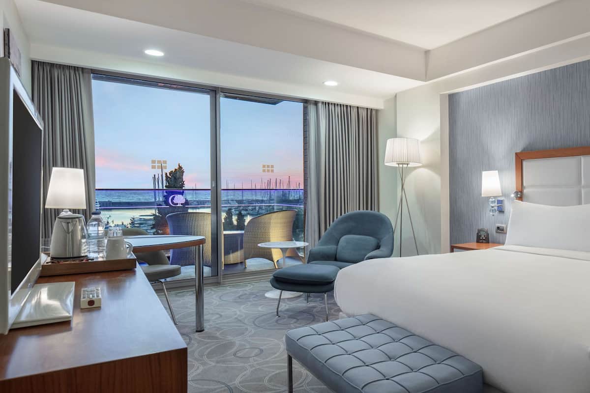 Habitación de hotel con una cama grande, un escritorio con lámpara, una silla, una mesita y vistas al mar a través de las ventanas.