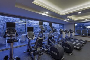 A gym with treadmills, elliptical machines, and stationary bikes, large windows, and a stone wall.
