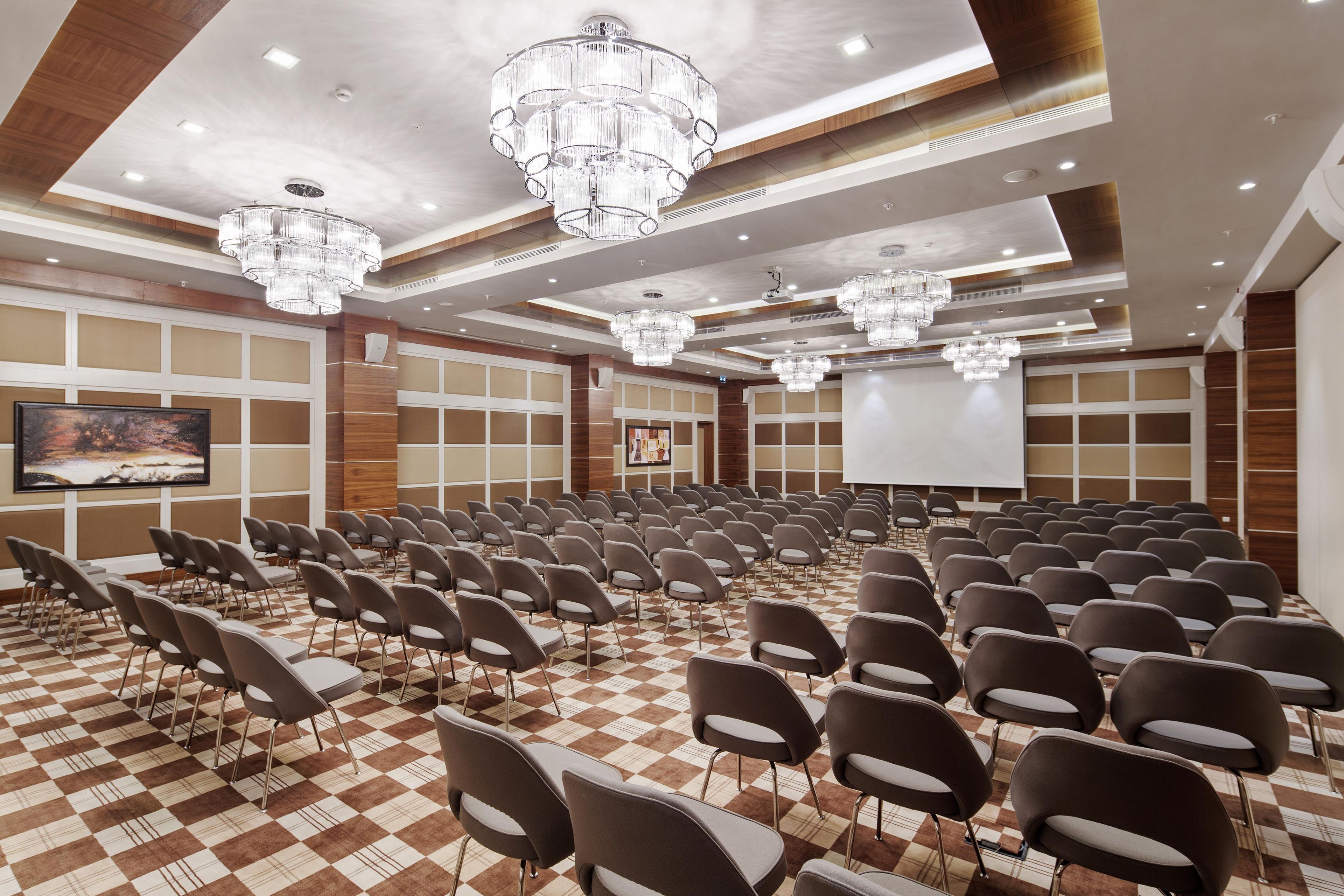 A conference room with rows of chairs, a large screen, and multiple chandeliers.
