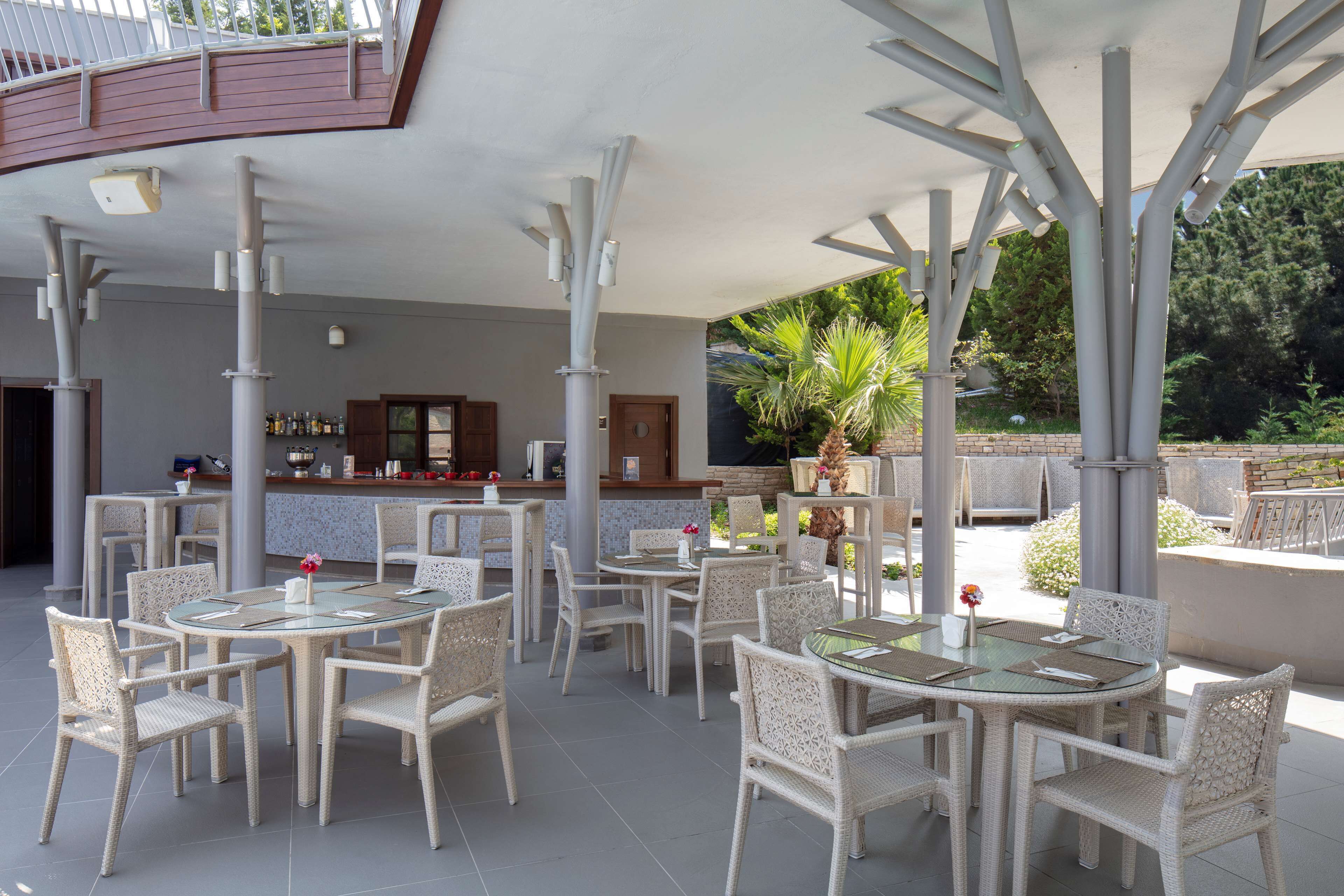 Un patio al aire libre con mesas redondas y sillas de mimbre, una zona de bar al fondo y vistas a la vegetación.