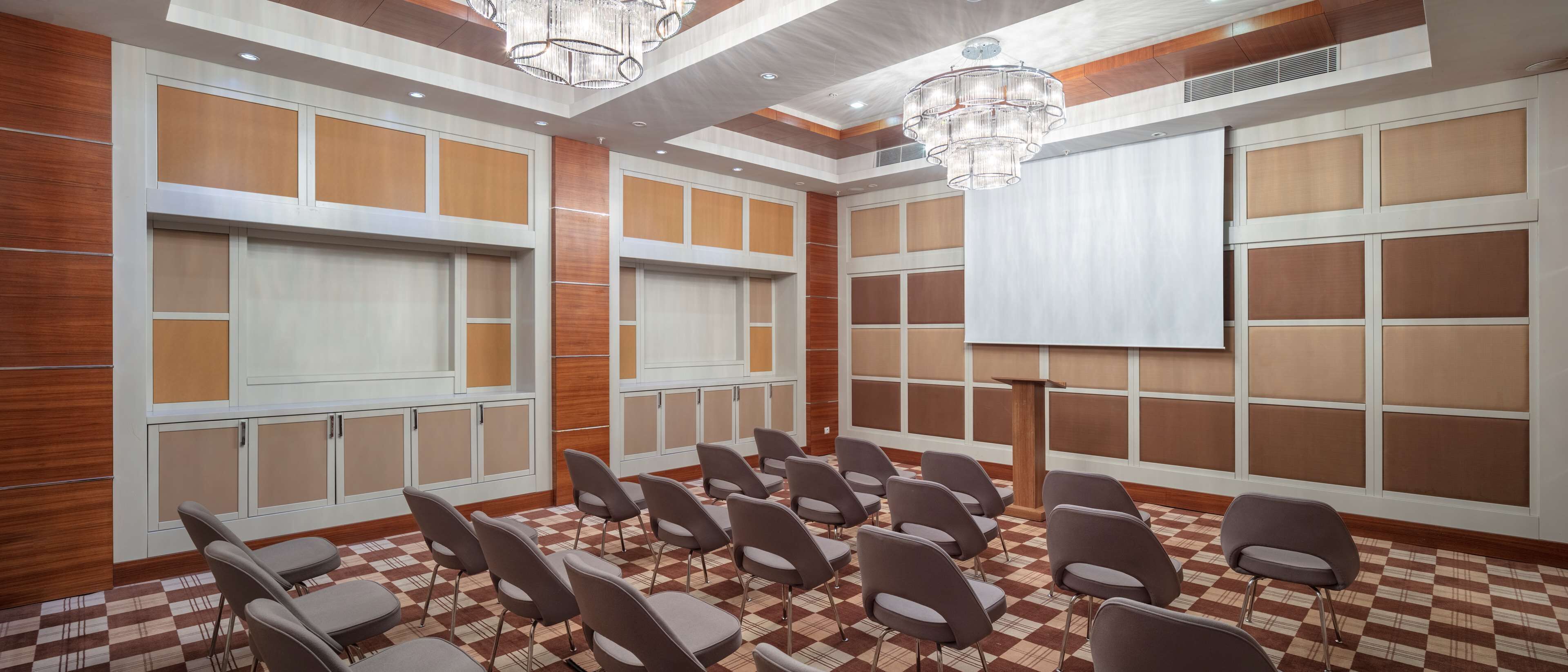 Un salón de conferencias con piso a cuadros, sillas grises, paneles de madera y una pantalla grande.