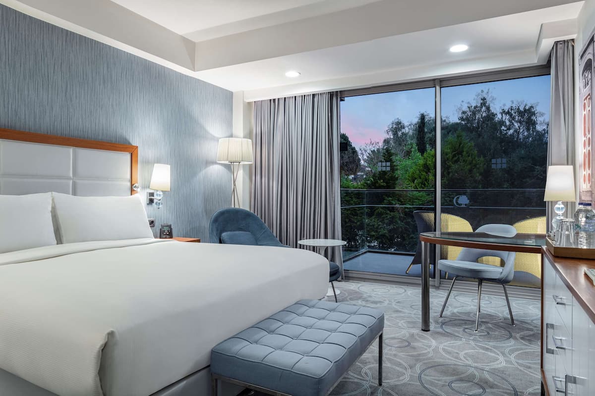 Habitación de hotel con una cama grande, un escritorio con computadora, una pequeña mesa con sillas y un balcón con vista a árboles y edificios.