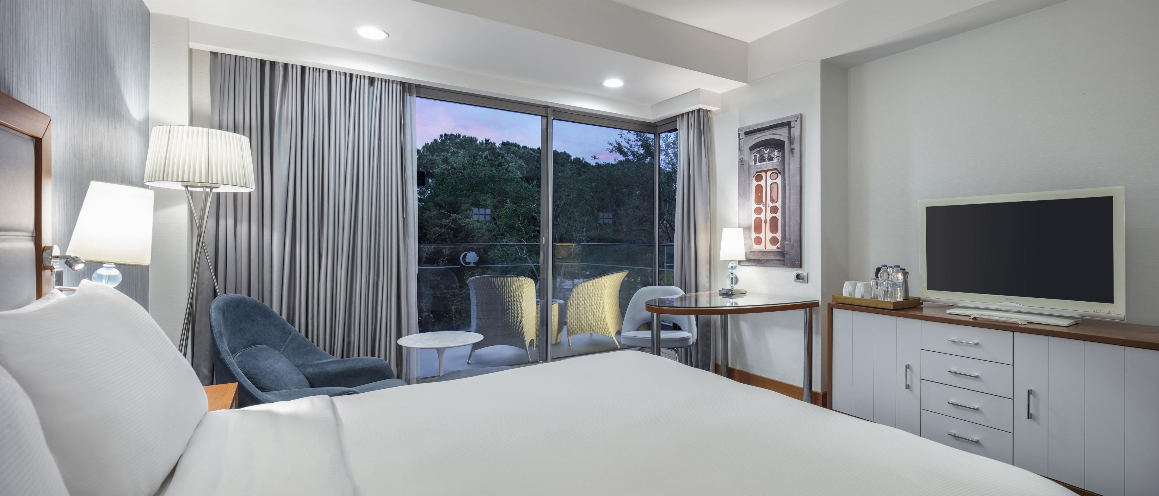 A hotel room with a bed, a TV, a desk with a lamp, and a balcony with chairs.