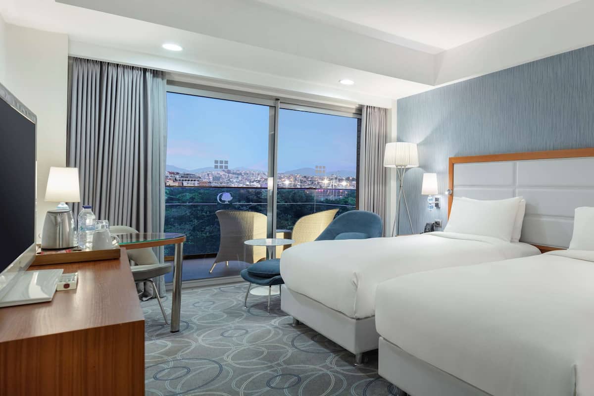 Habitación de hotel con dos camas, un escritorio, una silla y vistas a un paisaje urbano a través de amplios ventanales.