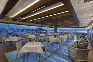 A restaurant with tables and chairs arranged on a glass floor, overlooking a cityscape. - Doubletree By Hilton Kusadasi - Special Class (Kusadasi)