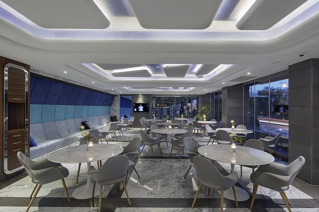 Un ristorante moderno con tavoli rotondi e sedie grigie, un'area salotto imbottita blu e vista sulla città attraverso ampie finestre.
