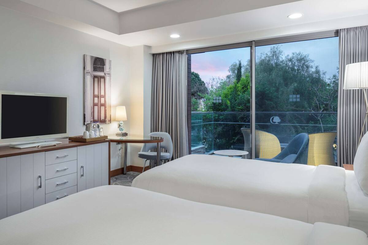 Habitación de hotel con dos camas, un televisor, un escritorio con lámpara y vistas a zonas verdes a través de amplios ventanales.
