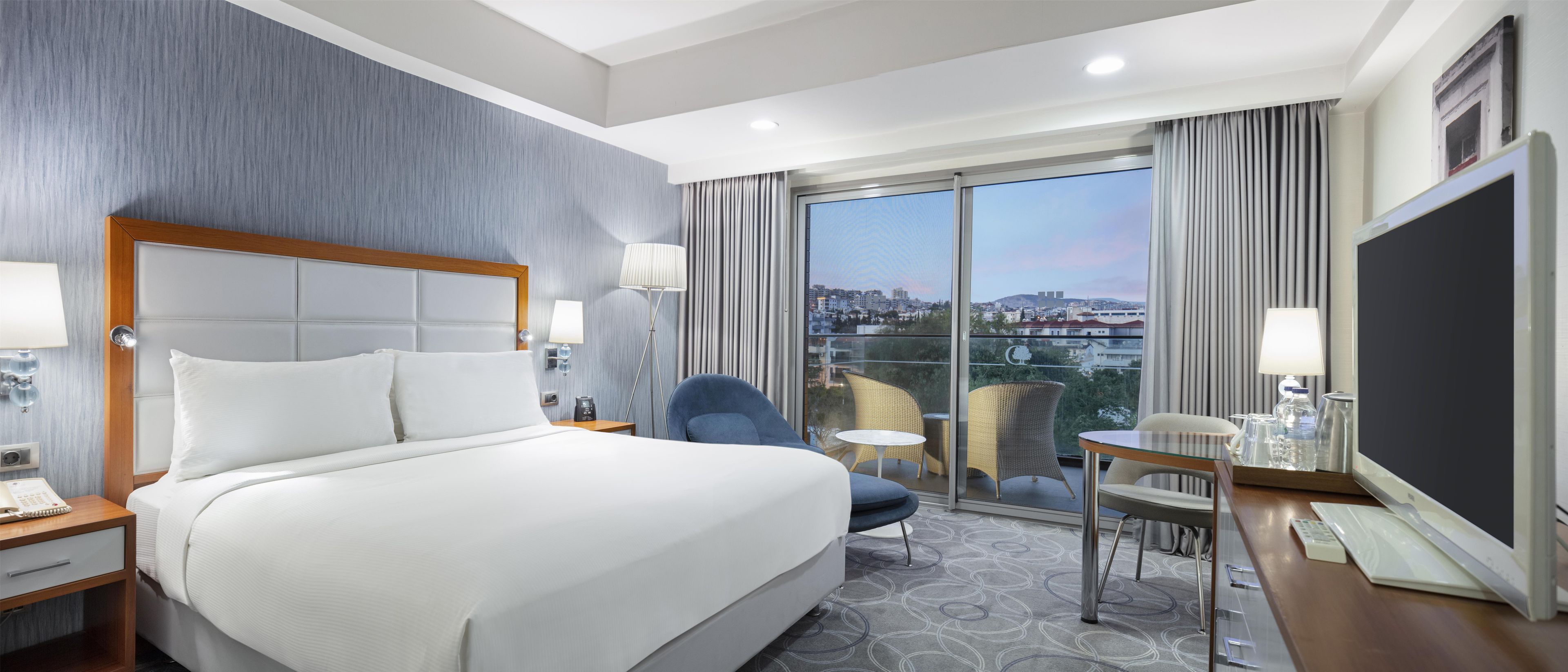A hotel room with a large bed, a desk, a chair, and a view of the city through a large window.