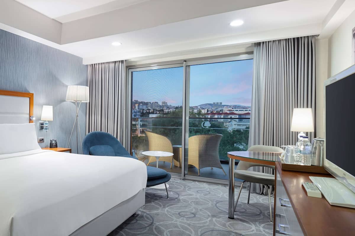 Habitación de hotel con una cama grande, un escritorio con una computadora, una silla y vistas a la ciudad a través de un ventanal amplio.