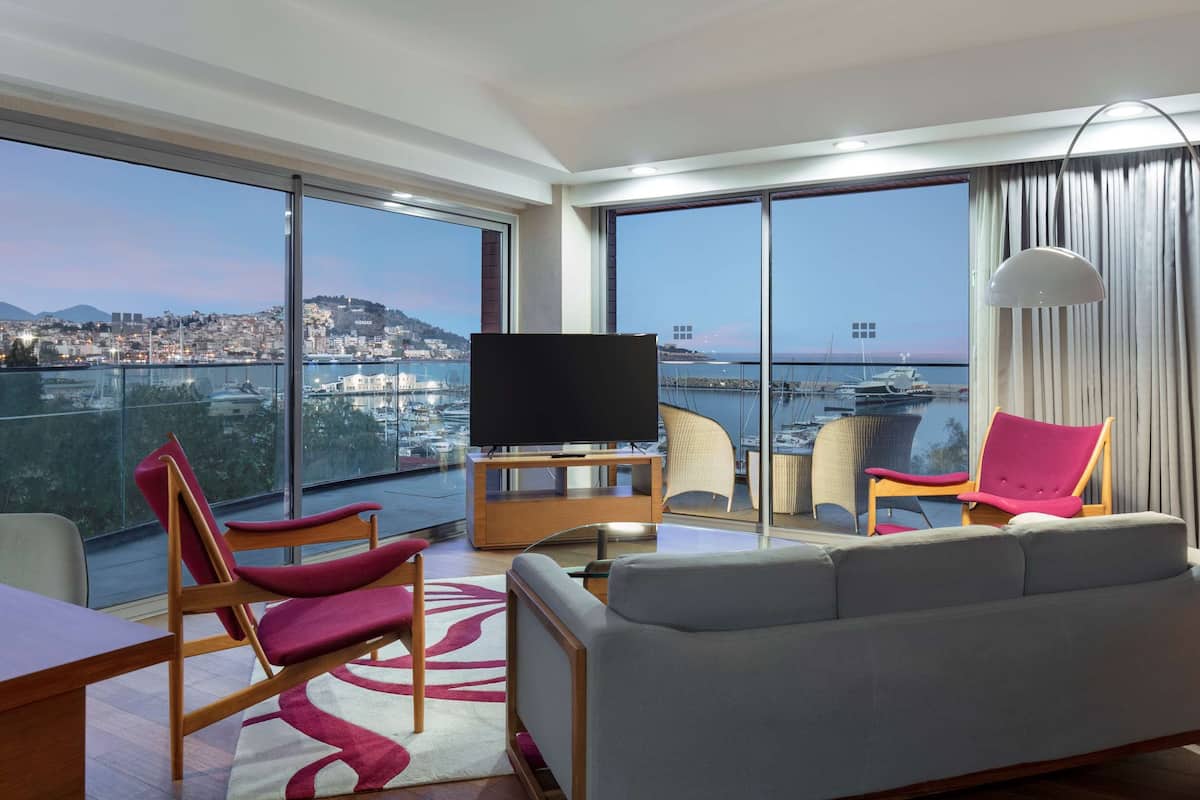 Una habitación de hotel moderna con un balcón grande, un televisor de pantalla plana, un sofá gris y un comedor.