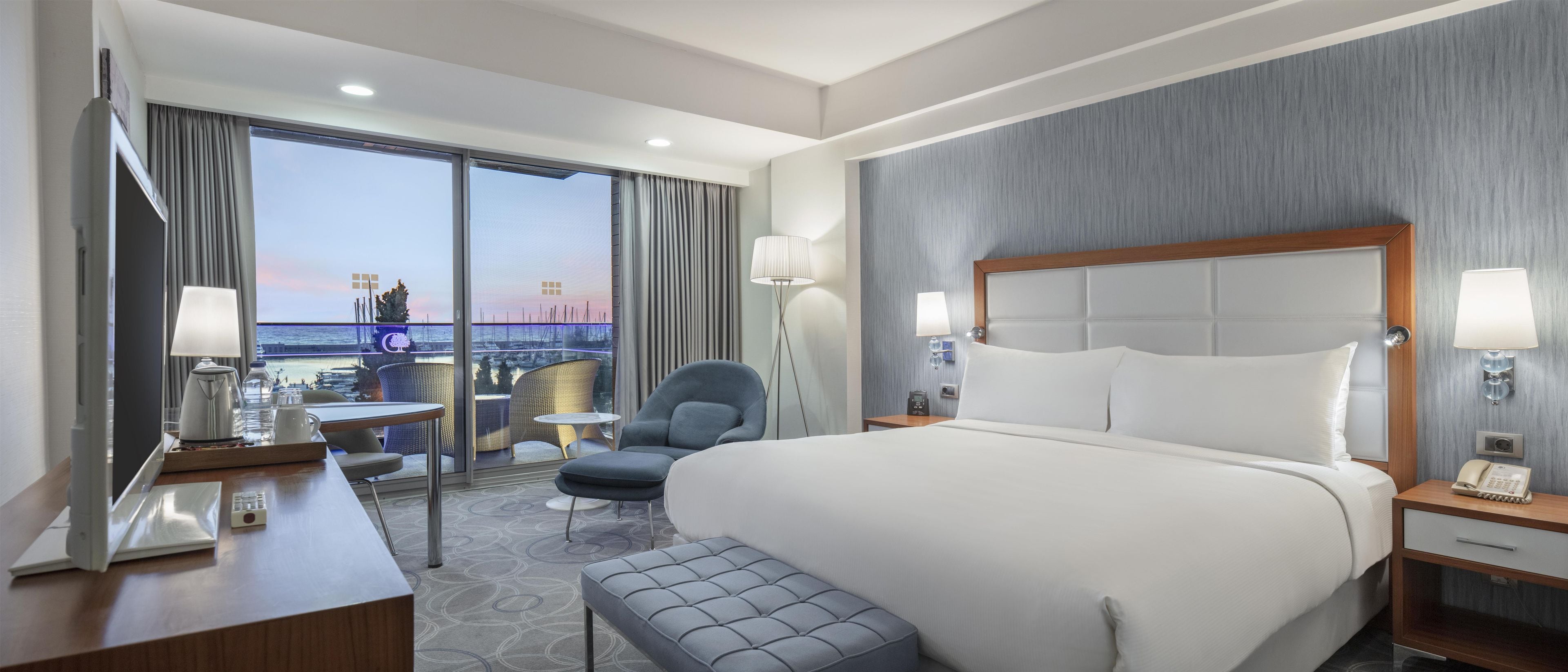 A hotel room with a large bed, a desk with a telephone, a chair, a small table, and a view of the sea through the window.