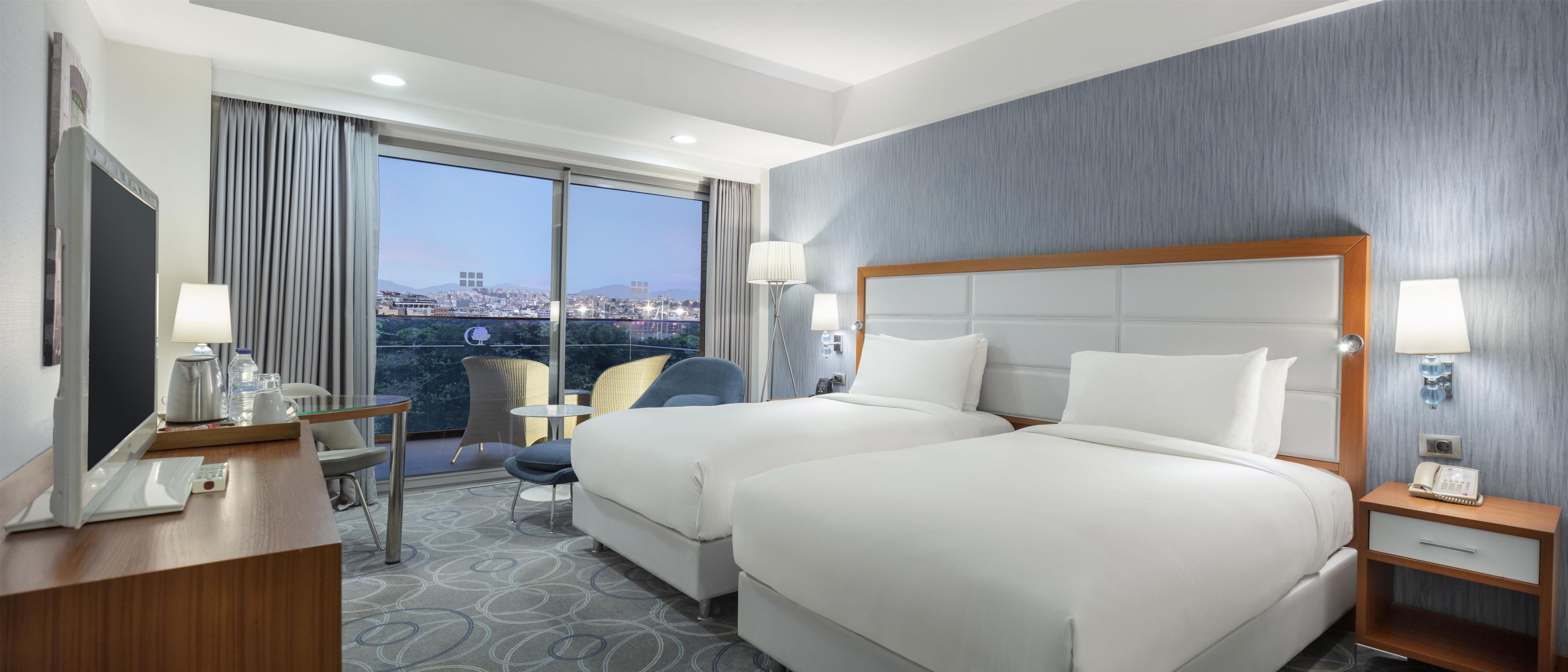 A hotel room with two beds, a desk, a chair, a small table, and a view of the city.