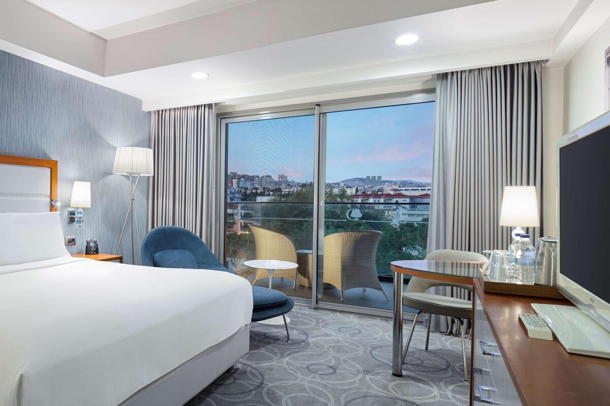 A hotel room with a large bed, a desk with a computer, a chair, and a view of the city through a large window.