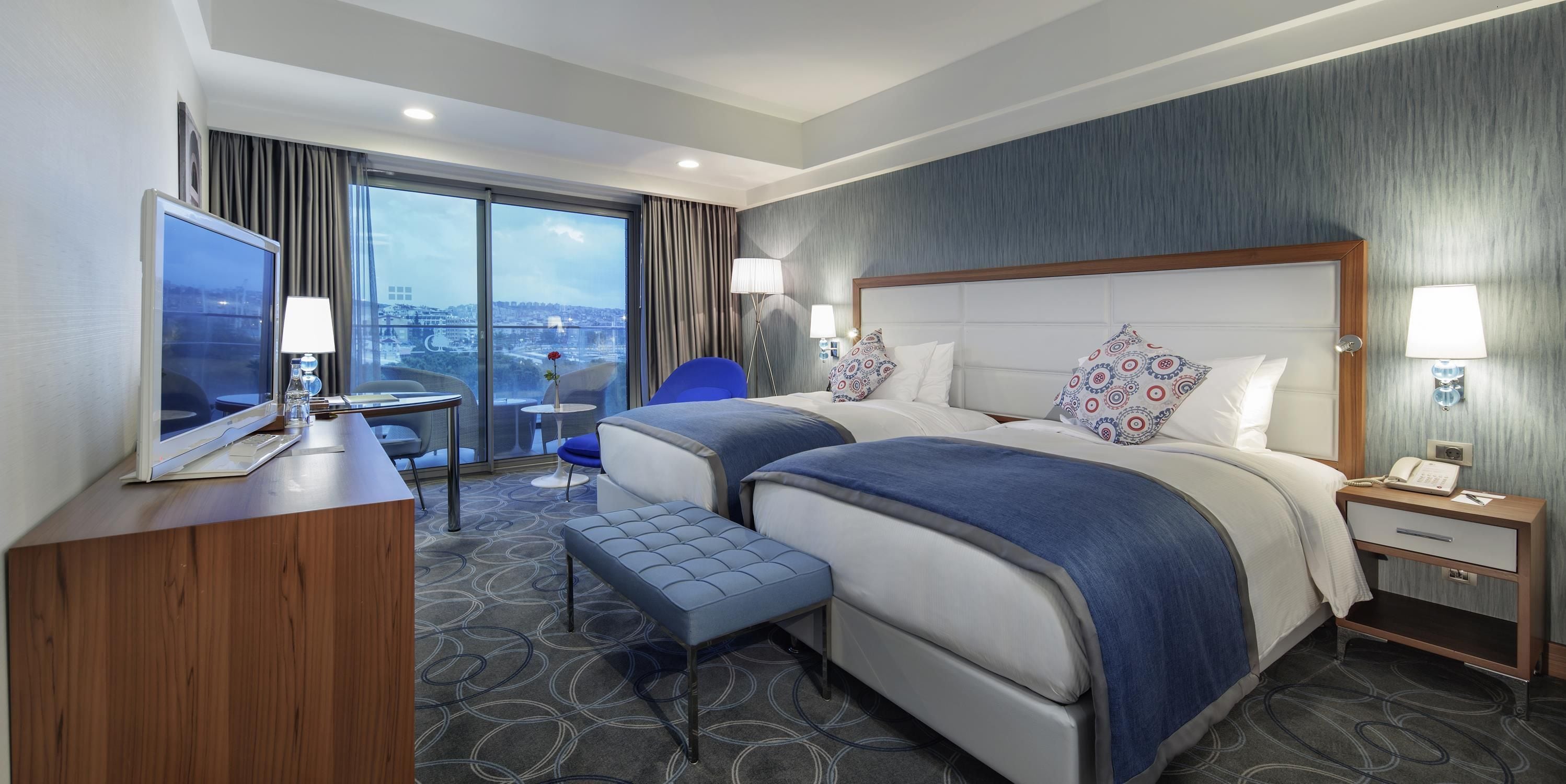 Habitación de hotel con una cama grande, un escritorio, una silla, una mesita y vistas a la ciudad.