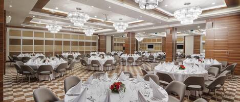 Un salón de banquetes con mesas redondas dispuestas para comer, sillas y candelabros de cristal.