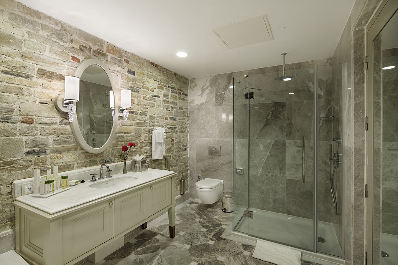 Un bagno con parete in pietra, un ampio specchio, un mobile bagno doppio, un WC e una cabina doccia in vetro.