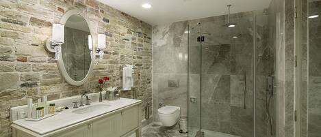 Un baño con pared de piedra, un espejo grande, tocador doble, inodoro y ducha con mampara de vidrio.